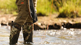 Angler's legs in stream wearing camouflage waders and First Lite pants, net and orange fly line
