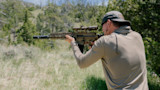 Man wearing baseball cap and earbuds aiming a scoped rifle with suppressor in grassy field near trees