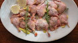 Raw quail on white platter with rosemary, thyme, lemon peel, halved garlic and coarse salt