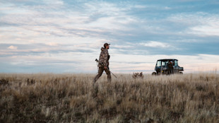 Hunter walking across dry grass carrying a rifle, UTV with passenger behind under big sky