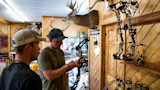 Two men examine a compound bow in a wood-paneled archery shop, mounted deer heads and sign "FRED BEAR"