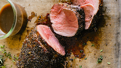 Sliced pepper-crusted roast with jar of gravy and thyme on wooden cutting board