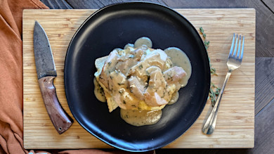 Sliced pheasant with herb gravy on toast on black plate, knife and fork on bamboo cutting board