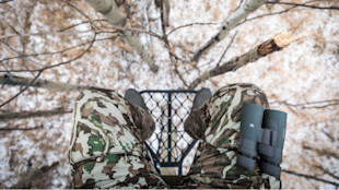 View from tree stand: camo-clad legs and binoculars over snowy forest floor