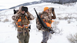 Two hunters using binoculars with chest harnesses in snowy field, camo and orange vests, rifle slung