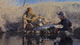 Two hunters kneeling by icy stream with beige cooler on frosty bank