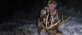Man kneeling in snowy woods at night holding a large buck's antlers