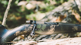 Camouflage bolt-action rifle with fluted bolt and scope resting on a log