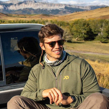 Man in sunglasses wearing First Lite hoodie sitting on truck tailgate with mountains behind