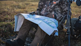 Hunter seated holding a topographic map; camo First Lite jacket, tripod and pack beside him