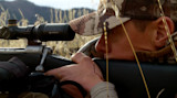 Hunter lying prone aiming rifle with Vortex scope through dry grass
