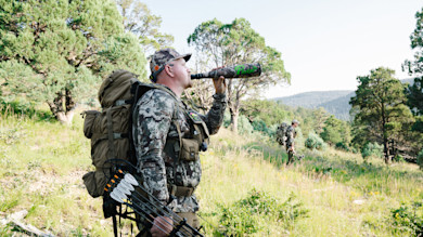 Hunter calling elk with Phelps elk call, carrying compound bow and backpack on grassy hillside