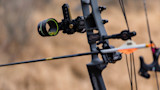 Compound bow with nocked arrow and sight, rain droplets on gear in tall grass