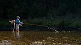 Man and girl fly-fishing in shallow river; man casting a long fly line over sunlit water