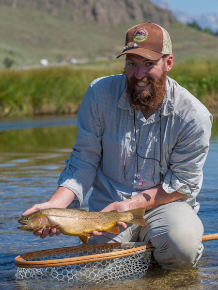 Angler kneeling in shallow river holding brown trout over wooden landing net