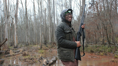 Smiling hunter wearing glasses and headlamp holds shotgun in flooded, leafless woods