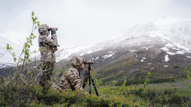 Two hunters in camouflage—one with binoculars, one using a spotting scope, snow‑capped mountains