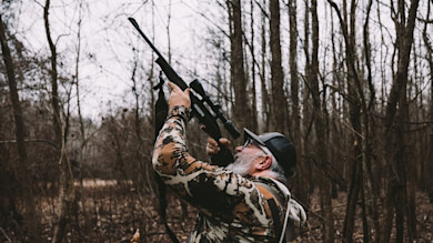 Hunter in camouflage aiming scoped rifle upward in leafless woods, wearing cap and glasses
