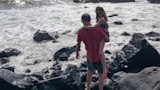 Man carrying girl on rocky shoreline as waves crash around them