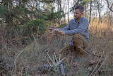 Man kneeling in woods holding deer antlers; another antler rack and a call lie on the ground