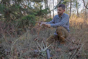 Man kneeling in woods holding deer antlers; another antler rack and a call lie on the ground