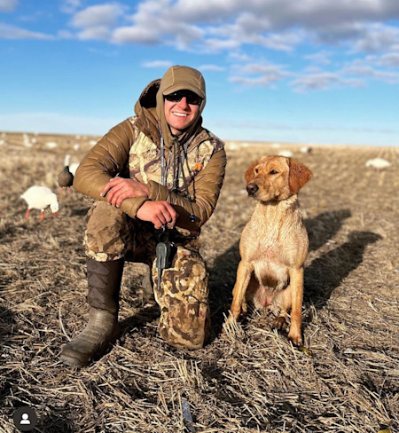 Hunter kneeling in harvested field beside wet golden retriever and white decoy geese