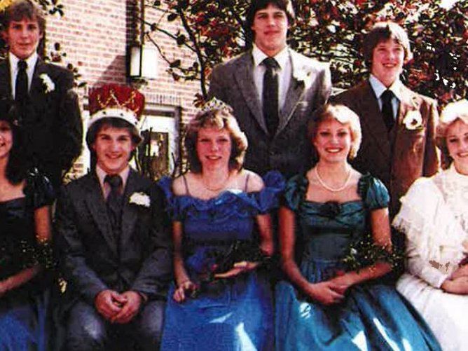 Teens in formalwear sitting and standing; two girls in blue gowns, seated boy wearing crown
