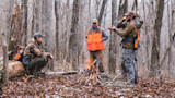 Three hunters in camo and orange vests gathered around a small campfire in a leafless forest