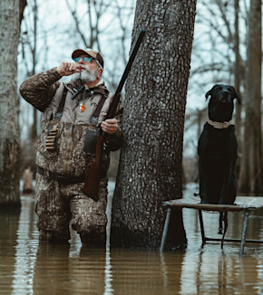 Hunter in camouflage waders blowing a duck call, holding shotgun; black lab seated on a blind