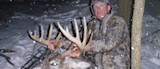Hunter in camouflage holding large buck antlers in falling snow, rifle nearby