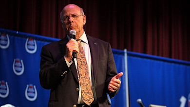 Man in suit and paisley tie speaking into microphone on stage with blue banners