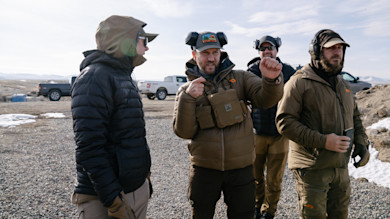 Clay Newcomb gesturing with ear protection beside three men at an outdoor shooting range