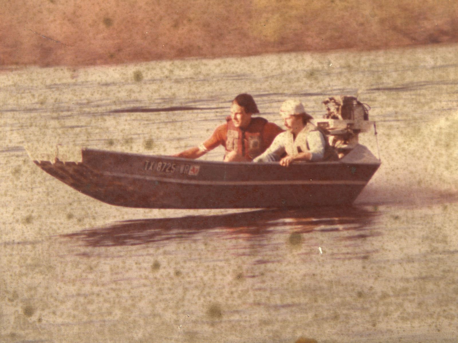 Two men in a small motorboat speeding across a lake; hull text "TX 8725 TR" visible