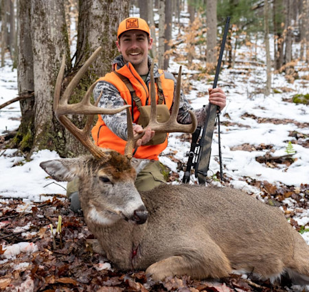Hunter kneeling in snowy woods holding rifle and large buck by its antlers, wearing orange vest and cap