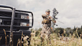 Hunter in camouflage holding compound bow beside pickup truck in grassy field