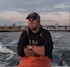 Man in orange bibs and black Huk hoodie on boat, bridge and water at dusk