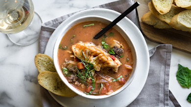 Cioppino with walleye and venison in white bowl, herb garnish, toasted baguette and white wine