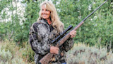 Woman in camouflage jacket holding leopard-print rifle with scope, smiling in grassy field