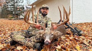 Hunter kneeling behind large whitetail buck, holding massive antlers; camo gear and bow