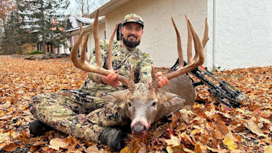 Hunter kneeling behind large whitetail buck, holding massive antlers; camo gear and bow
