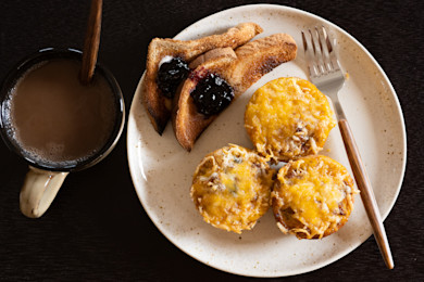 Three cheesy egg muffins on a plate with jam-topped toast triangles, fork, and mug of coffee