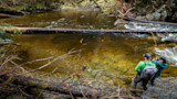 Two anglers wading in a clear forest stream, one casting a bent fly rod