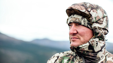 Hunter in camouflage hood and beanie, side profile looking toward distant mountains
