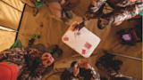 Four hunters in camouflage playing cards on a white cooler inside a tent