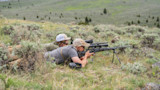 Two hunters lying prone in sagebrush; one aims a scoped rifle on a bipod, the other spotting