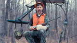 Hunter in orange vest and cap sitting in tree stand holding a shotgun, arm in cast