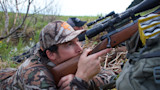 Hunter prone aiming a scoped rifle; second hunter lying behind in grass