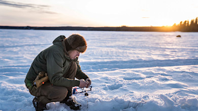 Man kneeling on frozen lake at sunset setting an ice-fishing tip-up beside a drilled hole