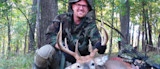 Hunter kneeling in wooded area holding buck's antlers with bow and arrows nearby