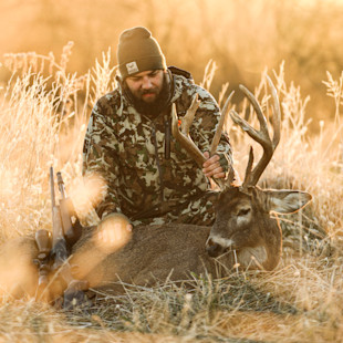 Hunter in camouflage wearing MeatEater beanie, kneeling beside large buck with rifle, holding antlers